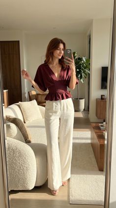 Woman in a maroon top and white pants taking a mirror selfie indoors