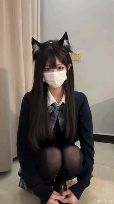 Person in a school uniform with cat ears and a face mask, squatting indoors