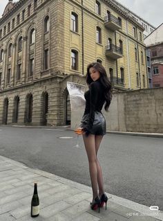 Woman in a black top and leather skirt holding a newspaper and a drink on a city street