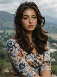Woman with long brown hair in a floral top outdoors with mountains in the background