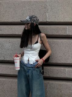 Person in a camo hat and baggy jeans holding a drink against a stone wall
