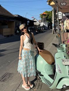Person with long hair, bandana, and sunglasses standing next to a scooter on a street