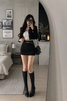 Woman in a black cropped jacket, mini skirt, and knee-high boots taking a mirror selfie