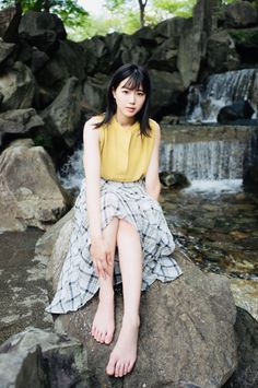 Woman with black hair in a yellow top and plaid skirt sitting barefoot by a waterfall