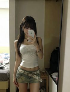 Woman in a white tank top and denim mini skirt taking a mirror selfie