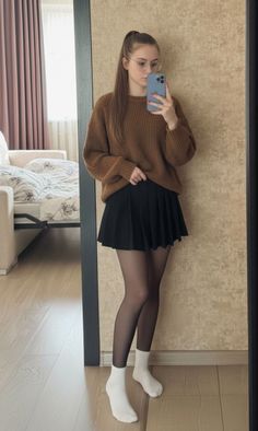Woman in a brown sweater and black pleated skirt taking a mirror selfie