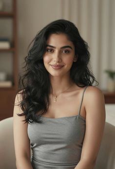Woman with long black hair wearing a gray camisole and gold necklace, smiling indoors