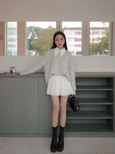 Woman in a grey sweater and white pleated skirt standing indoors