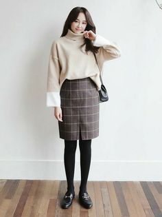 Woman in a beige turtleneck sweater and plaid skirt standing indoors
