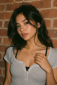 Woman with long dark hair wearing a ribbed top and gold necklace against a brick wall