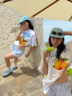 Two women in cropped t-shirts and shorts on a beach with an umbrella
