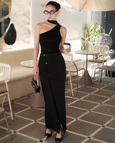 Woman in a black one-shoulder top and long skirt holding a coffee cup at an outdoor cafe