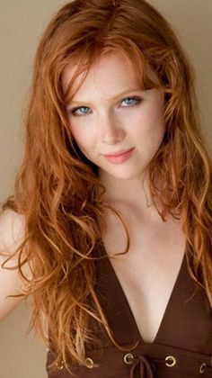Woman with long red wavy hair and blue eyes wearing a brown top