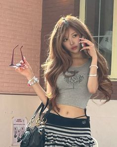 Woman with long hair wearing a crop top and striped skirt, holding sunglasses and a phone