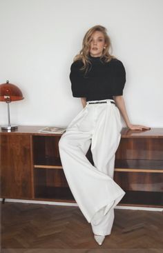 Woman with long blonde hair wearing a black top and white wide-leg pants indoors