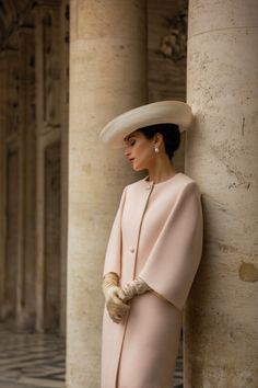 Woman in a pink coat and wide brim hat leaning against a column
