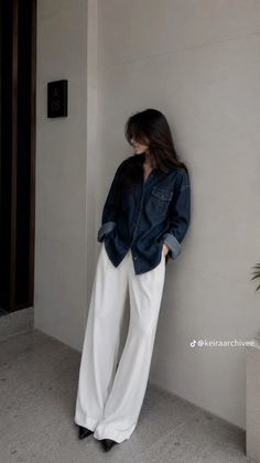 Woman in a denim shirt and white pants leaning against a wall indoors