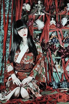 Woman in a red kimono with long black hair and red eyes, surrounded by Japanese dolls