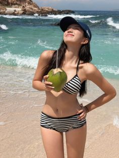 Woman in a striped bikini and baseball cap holding a coconut drink on a beach