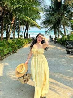 Woman in a yellow dress holding a straw hat on a path lined with palm trees near the beach