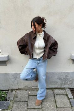Woman in a brown leather jacket and light wash jeans leaning against a wall
