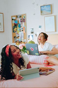 Two young women laughing and relaxing on a bed with a laptop and tablet