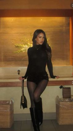 Woman in a black top, sequin shorts, and tall boots holding a handbag indoors