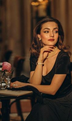 Woman with long wavy hair and pearl necklace in a black top, sitting indoors