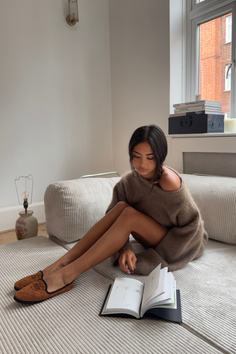 Woman in an oversized brown sweater and slippers sitting on a ribbed sofa indoors