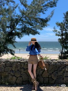 Woman in a straw hat and blue shirt standing by a stone wall at the beach