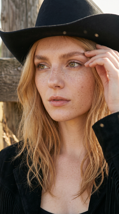 Woman with blonde hair and freckles wearing a black cowboy hat and jacket outdoors