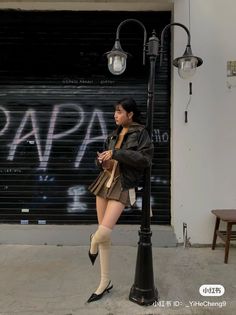 Woman in a leather jacket and pleated skirt leaning on a street lamp