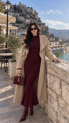 Woman in a burgundy dress and long coat posing on a stone wall overlooking a city