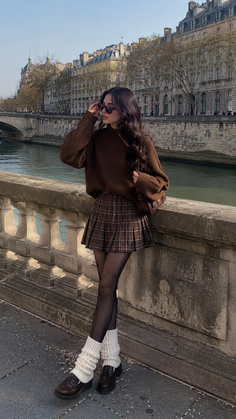 Woman in a brown sweater and plaid skirt with leg warmers on a bridge overlooking a city