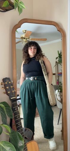Woman with curly hair in a black top and green pants taking a mirror selfie