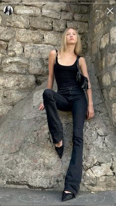 Blonde woman in a black tank top and jeans leaning against a stone wall