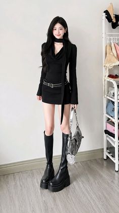 Woman in a black dress and knee-high boots holding a handbag indoors