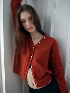 Woman in a red cardigan and black bra leaning against a white wall