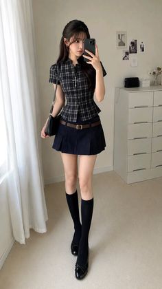 Woman in a plaid top and mini skirt taking a mirror selfie indoors