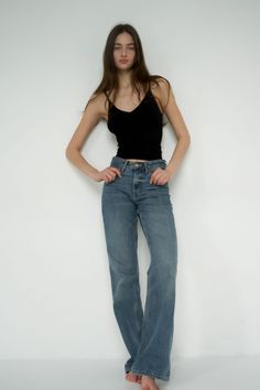 Woman with long hair wearing a black camisole and blue jeans against a white wall