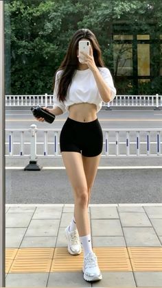 Woman in a white crop top and black shorts taking a mirror selfie outdoors