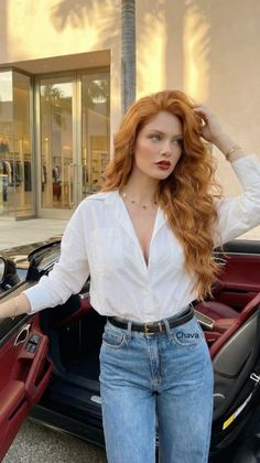 Woman with red hair in a white shirt and blue jeans next to a car