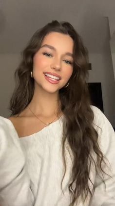Woman with long wavy hair wearing a white top and gold jewelry, smiling