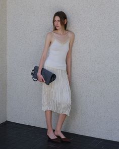 Woman in a white camisole and pleated skirt holding a gray bag outdoors