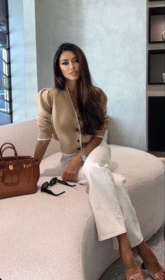 Woman with long dark hair wearing a cropped cardigan and white pants, sitting indoors