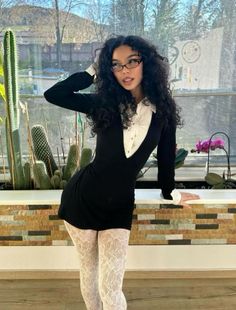 Woman with curly hair and glasses wearing a black dress and white lace tights indoors