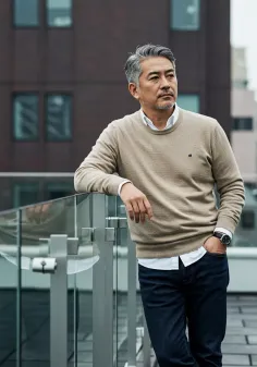 Man with gray hair in a beige sweater and jeans leaning on a railing outdoors