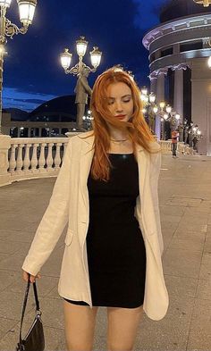 Woman with long red hair in a black dress and white blazer on a city street at night
