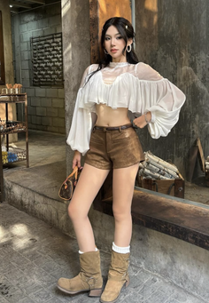 Woman in a white cropped blouse and brown shorts with cowboy boots indoors