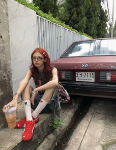 Person with red hair and glasses sitting on a curb next to a car